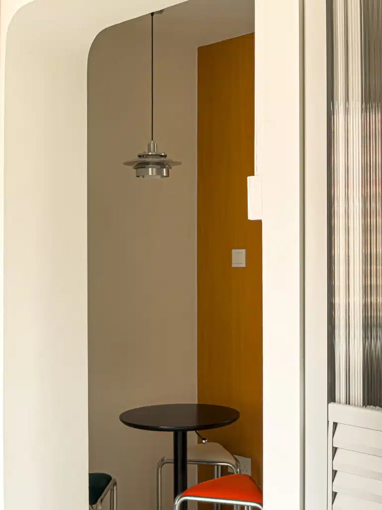 Small nook featuring a circular black table, pendant lamp, and colourful retro stools. The Louvre entrance door of common bathroom is in view, its top half is made of flutted glass finishes.