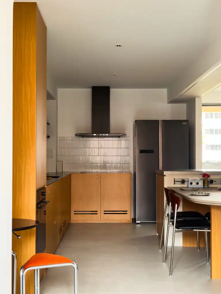 Full view of a Bauhaus-inspired kitchen featuring warm wood cabinetry, a tiled backsplash, and a central island with contrasting grey countertop. The clean geometry and tonal balance define the open-concept layout.