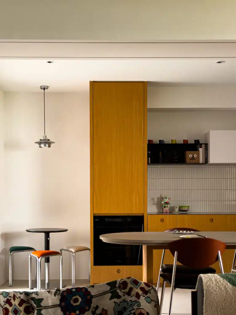 View from the living room looking into the kitchen, showing how the open layout connects both spaces. The yellow wood cabinetry contrasts with darker shelving and white upper cabinets for visual layering.