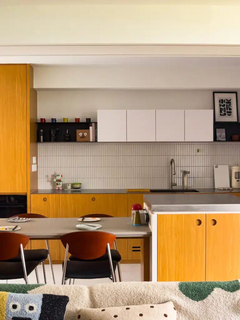 Front-on view of a Bauhaus-inspired open kitchen seen from the living room: warm oak cabinetry below crisp white uppers, a fine vertical-tile backsplash, and a long black shelf with colourful cups and framed art. A grey peninsula is set with plates and a small red-topped “mushroom” lamp; a hint of the sofa/throw anchors the foreground.