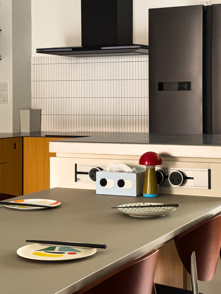 Close-up of the kitchen island with graphic tableware, a mushroom lamp, and built-in sockets. Playful colors sit against a muted grey counter, showing the home’s mix of function and character.