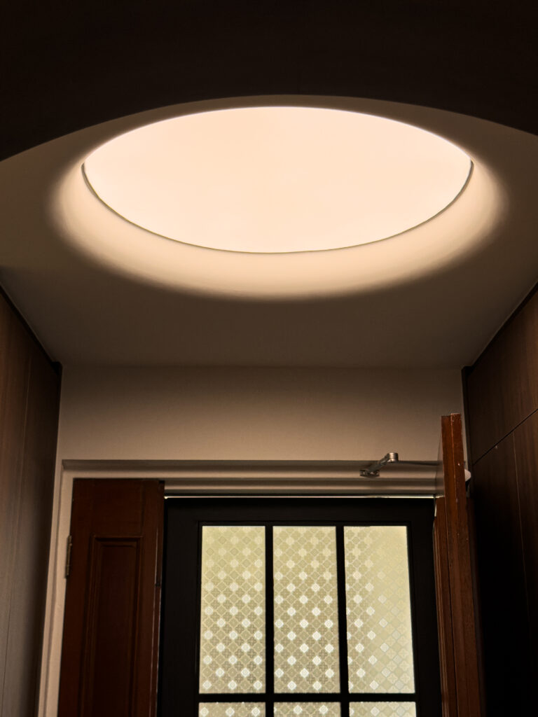 Soft ambient light radiating from a round recessed dome ceiling, diffused through a translucent fabric light. The gentle curvature of the microcement ceiling is framed by warm-toned wall panels and a partially opened wooden door below.