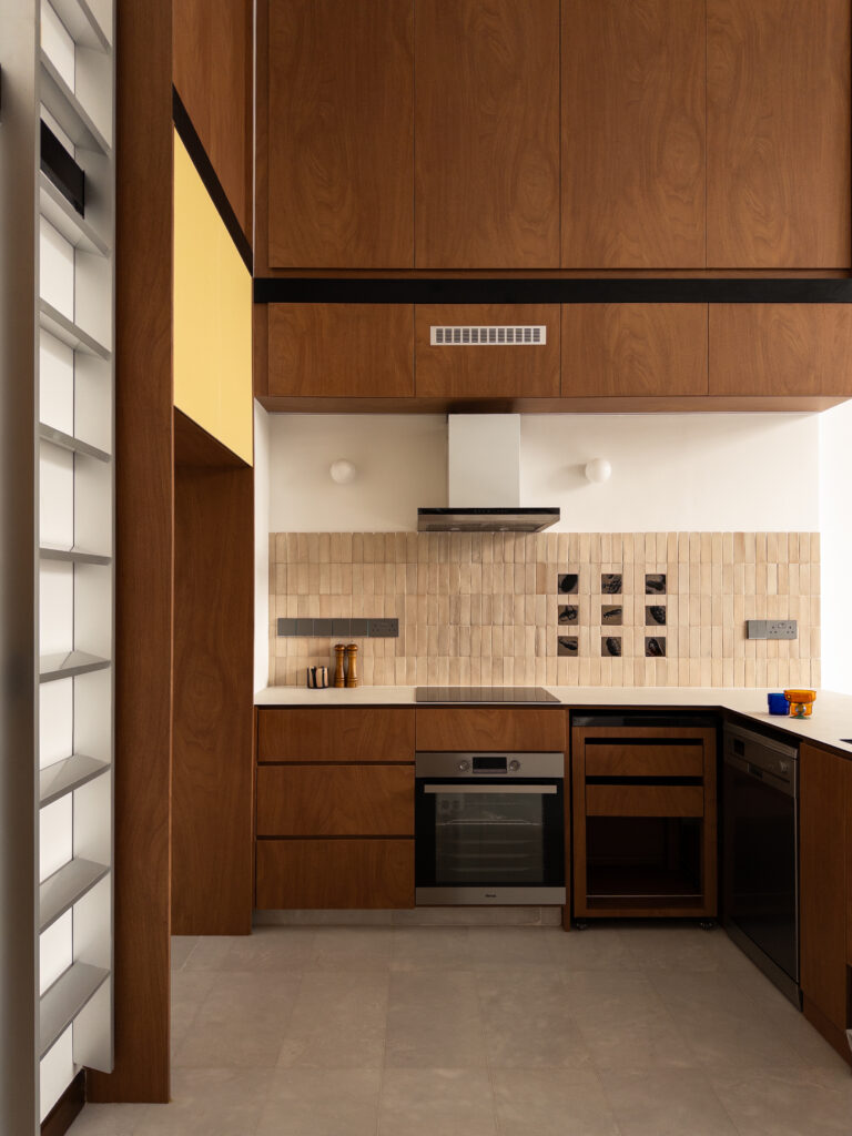 Cooking area with tall walnut cabinets, textured beige backsplash tiles, light countertop, and built-in oven in a double-volume kitchen.